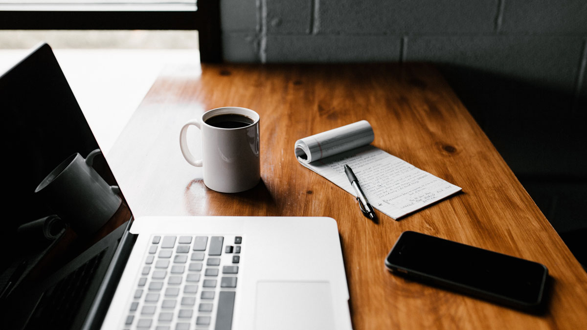 A laptop, white mug, notepad, pen and phone sitting on a wooden desk